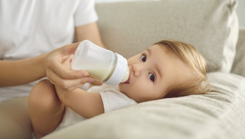 Baby enjoying nutritious meal. Mother feeding baby on sofa. Nanny formula feeding little child as alternative to breastfeeding. Baby boy or girl lying on couch and drinking milk from plastic bottle