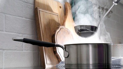 Adding water with ladle into steaming saucepan on stove in kitchen