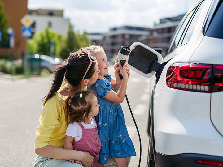 A mother with two young daughters helping one of them plug in an electric vehicle with a charger in a street charging port