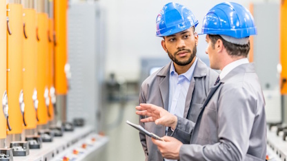 Deux professionnels en environnement industriel examinent une tablette devant des armoires électriques et équipements techniques. La scène illustre un échange autour de la performance, de la sécurité ou de la conformité des installations.