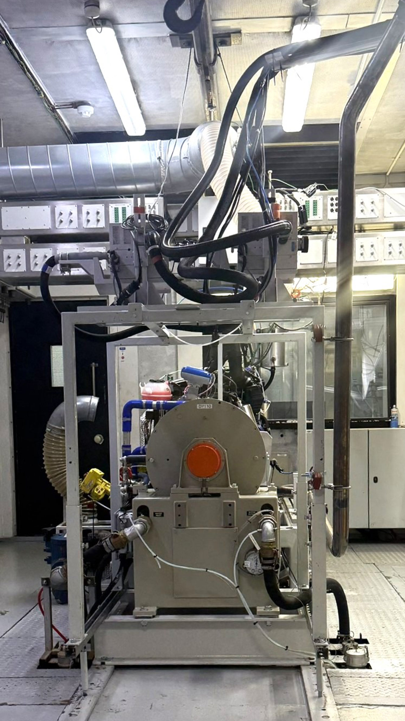 A front view of an automotive engine test cell showing a mounted engine connected to a large dynamometer. Overhead cables and ventilation ducts run across the ceiling, with control panels and industrial equipment lining the walls in a clean, enclosed test environment.